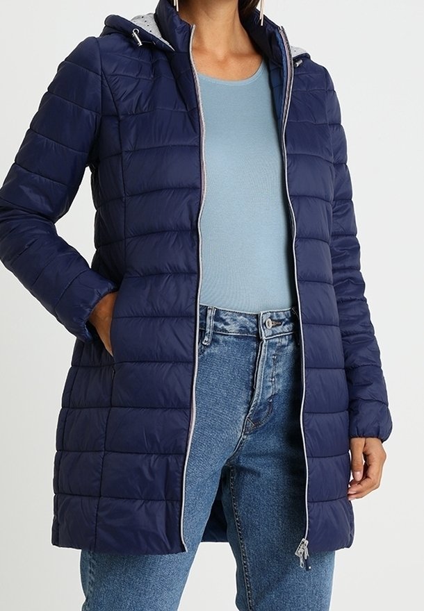 Abrigo acolchado azul marino con capucha ajustable, diseño acolchado, cremallera gris y bolsillos laterales. Lleva puesto encima una camiseta azul claro y pantalones vaqueros.