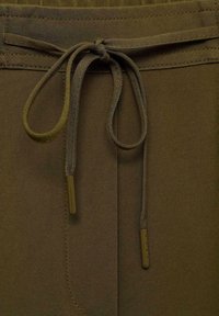 Un pantalon en tissu marron présente une taille à cordon, avec des liens fins et lisses et de petites agrafes métalliques. La texture est douce et légèrement côtelée.