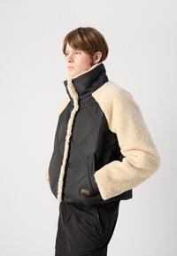 Πουφ jacket με μαύρο νάιλον σώμα και μπεζ ψεύτικη γούνα στα μανίκια. Διαθέτει ψηλό γιακά, διαμαντένιο σχεδιασμό και λεπτομέρεια με λογότυπο.