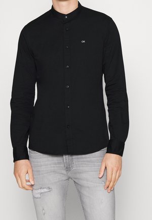 Mann trägt ein eng anliegendes schwarzes Hemd mit Mandarin-Kragen und "CK" Logo auf der Brust sowie hellgraue, abgenutzte Jeans.