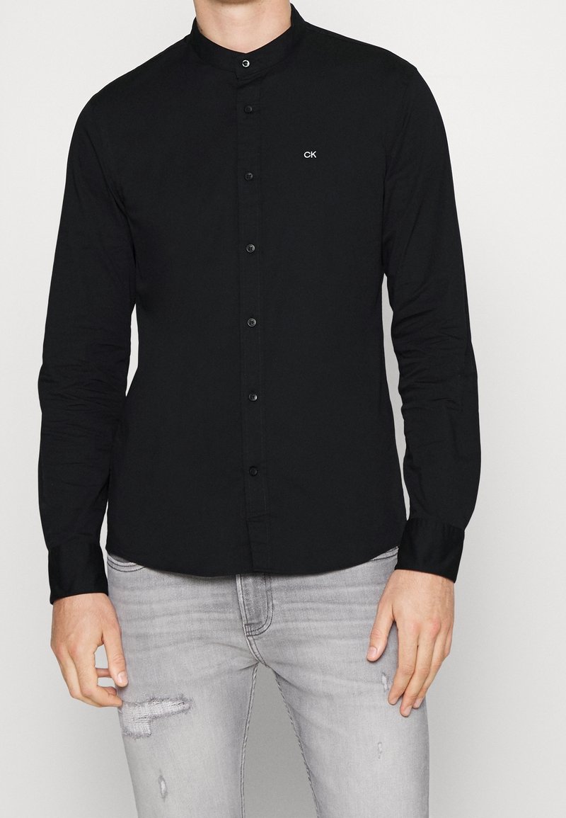 Homme portant une chemise noire slim à col mandarin avec logo « CK » sur la poitrine et un jean gris clair usé.