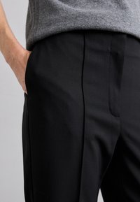 Fekete, sima textúrájú, személyre szabott nadrág, elülső redőkkel és oldalsó zsebekkel, derékban egy szürke kötött pulóverrel párosítva.