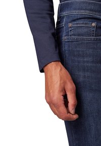 Chemise à manches longues bleu marine en tissu texturé avec un ourlet arrondi, associée à un jean en denim foncé avec deux poches arrière et des coutures discrètes.