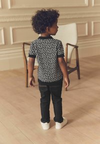 Schwarz-weiß gemustertes T-Shirt, kurze Ärmel, kombiniert mit dunklen Jeans und weißen Slippern. Steht auf hellem Holzboden.