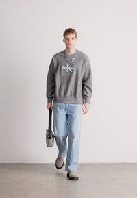 Šedá mikina s logem, světle modré volné džíny, šedé nazouvací boty a šedá kabelka přes rameno. Model má krátké vlasy a nosí náhrdelník.