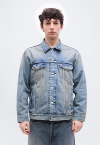 Νεαρός άνδρας με σγουρά μαλλιά, φορώντας ανοιχτό μπλε τζιν μπουφάν και τζιν παντελόνι, στέκεται μπροστά σε λευκό απλό φόντο.