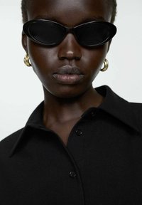 Occhiali da sole neri con un design angolare, grandi orecchini a cerchio dorati, e una camicia nera con bottoni, creando un aspetto elegante e moderno.