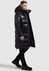 Abrigo largo y acolchado en un oscuro tono navy, con un alto cuello, acabado brillante, segmentos horizontales gruesos y bolsillos laterales con cremallera. Combinado con pantalones y botas negras.