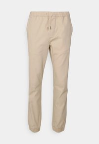 Pantalon beige à taille élastique avec cordon de serrage, jambes fuselées et poignets élastiques ; matière en coton lisse sans motifs ni accents visibles.