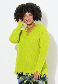 Pull en tricot vert citron avec un col en V, des bords effilochés et des manches longues. Porté par-dessus des leggings camouflés étoilés à motifs.