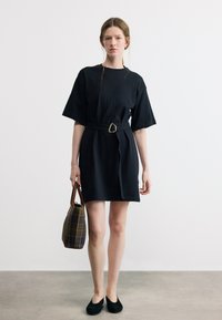 Νεαρή γυναίκα με μαύρο φόρεμα με ζώνη που κρατά καφέ καρό τσάντα, στέκεται μπροστά σε απλό λευκό φόντο με μαύρα παπούτσια.