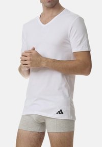 White short-sleeve v-neck t-shirt made of cotton, featuring a black Adidas logo on the lower left side. Worn with gray boxer shorts.