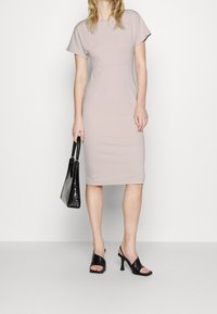 Tät passande klänning med kort ärm i ljus beige tyg; knälång design; slät textur. Accessoarer med en svart, strukturerad handväska och svarta klackar.