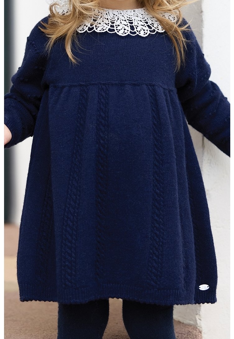 Tout-petit portant une robe en tricot bleu marine avec des motifs torsadés et un col en dentelle blanche, debout les bras légèrement écartés.