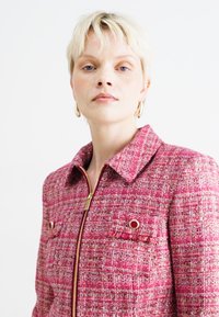 Vaate tweed-puku, vaaleanpunaisena, jossa on tekstuurinen pinta, kultainen vetoketju ja messinkiset napit rintataskuissa. Viistoreunainen kaulustyyli.