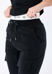 Pantaloni cargo neri con fascia elastica in vita e cordino regolabile. Presentano due tasche laterali e un polsino bianco con logo e testo nero.