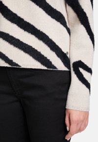 Crème gebreid sweater met zwarte zigzagstrepen, met een zachte, gestructureerde afwerking. Met een zilveren logodetail aan de zijkant, gedragen over zwarte broek.