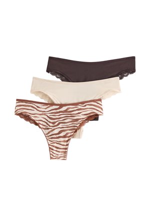 Donna in piedi scalza contro una parete bianca, indossa un reggiseno marrone scuro con rifiniture in pizzo e mutande a vita alta abbinate.