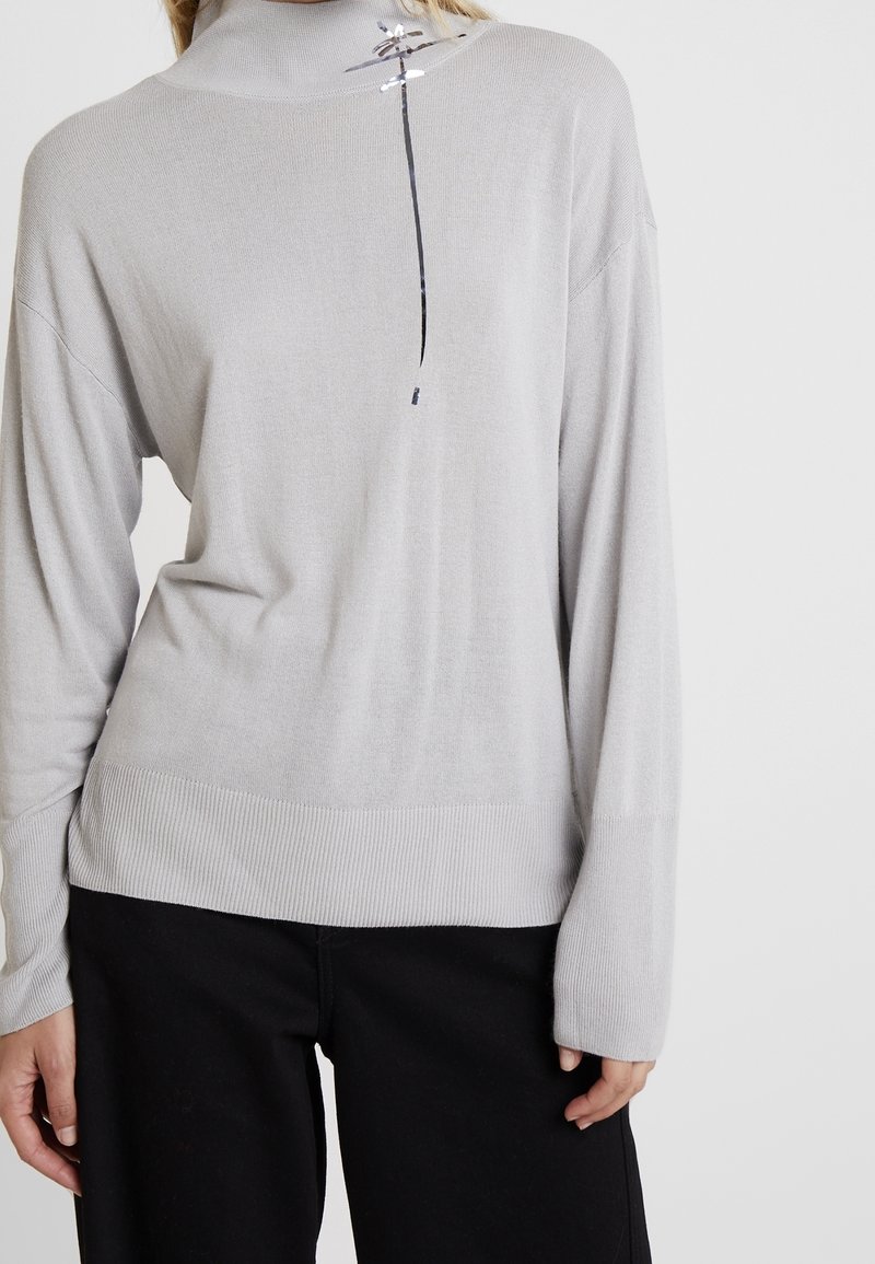 Ljusgrå, långärmad polotröja gjord av mjukt material, med ribbad fåll och en liten dekorativ detalj vid halsen.