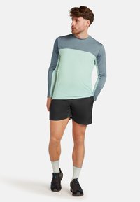 Långärmad träningsskjorta i teal och grått tyg, kombinerad med svarta shorts och mörka skor med ljusa accenter. Andningsbar och lätt design.