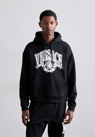 Jonged man in een zwarte Versace hoodie en bijpassende zwarte broek met het Medusa-logo, staand tegen een effen lichtgrijze achtergrond.