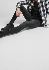 Persona sentada en el suelo, vistiendo una camisa de cuadros en blanco y negro, camiseta negra, jeans ajustados oscuros y zapatos negros, con anillos en la mano izquierda.