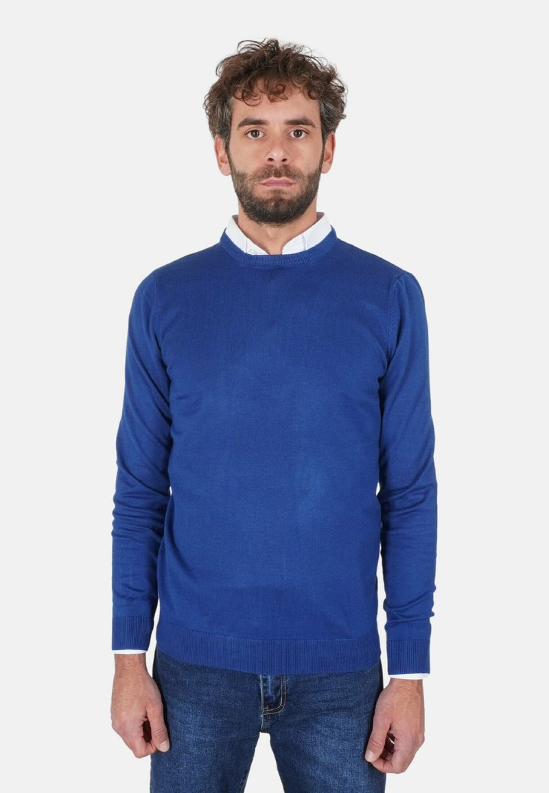 Maglione blu lavorato a maglia con scollo rotondo, polsini e orlo a coste. Indossato sopra una camicia bianca con colletto, abbinato a jeans blu.
