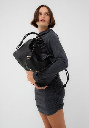 Femme portant une robe ajustée gris foncé, tenant un grand sac à main en cuir noir avec deux anses, sur un fond clair uni.