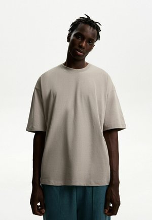 Light grey oversized t-shirt made of cotton with a round neckline and short sleeves. Model stands with hands at sides, wearing blue pants.