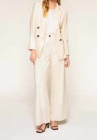 Blazer ajusté beige clair avec boutons noirs, associé à un pantalon large taille haute assorti. Tissu lisse et silhouette ajustée.