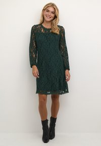 Robe en dentelle verte à manches longues, avec un col rond et une coupe décontractée. Tissu texturé avec des motifs floraux. Portée avec des bottines noires à la cheville.