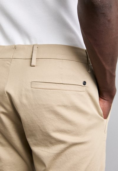Beige bomuldsbukser med en glat tekstur, der har en enkelt baglomme og et sort metalakcent. Designet med en lige pasform.