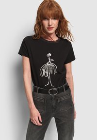 Mujer pelirroja vestida con una camiseta negra con un dibujo abstracto en blanco de un pájaro, metida en unos jeans gris oscuro con cinturón negro.