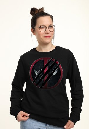 Kobieta w okularach, z włosami upiętymi w koka, ubrana w czarną bluzę z podartym logo Deadpool na białym tle.