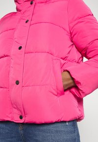 Ljusrosa puffjacka med svarta tryckknappar, sidofickor och en hand i den högra fickan, parat med blå jeans.