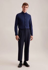 Marineblauw shirt met lange mouwen gemaakt van glad materiaal, voorzien van een knoopsluiting aan de voorkant en een klassieke kraag, gecombineerd met nauwsluitende donkere broek en zwarte schoenen.