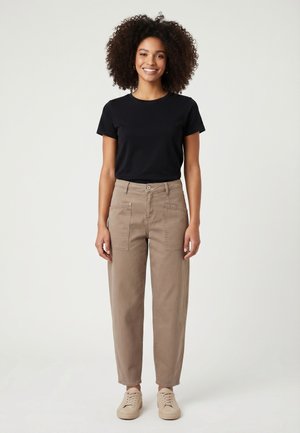 Jonge vrouw staand, draagt een zwart T-shirt met korte mouwen, beige hooggetailleerde broek met grote zakken en beige sneakers, glimlachend.