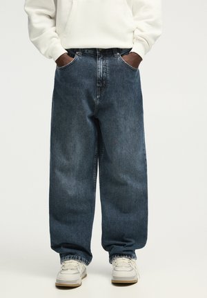 Blugi largi din denim albastru cu o textură estompată, cusături duble și fire de contrast, asortați cu un hanorac alb și adidași.