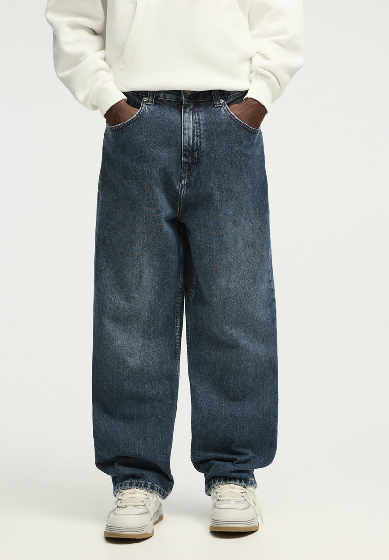 Løse blå denim jeans med en falmet tekstur, dobbeltstikninger og kontraststing, parret med en hvid hættetrøje og sneakers.