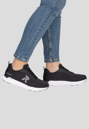 Personne portant des chaussures de sport noires avec des semelles blanches et un jean bleu sur un fond clair uni.
