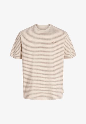 Blond jongetje staat en lacht, draagt een licht gestreept t-shirt, losse lichtblauwe spijkerbroek en bruine leren schoenen op een effen achtergrond.