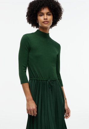 Woman with curly hair wearing a dark green, long-sleeve, high-neck pleated dress with a waist tie and gold bracelet on left wrist.