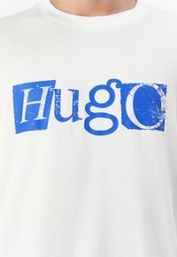 Camiseta de algodón blanca que presenta un gran texto distorsionado en azul que dice "HuGo" con diferentes estilos de fuente. Diseño simple y casual con gráficos impresos en serigrafía.