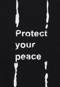 La tela de algodón negra presenta un texto blanco en negrita que dice "Protege tu paz", con acentos texturizados a ambos lados.