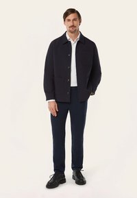 Marine casual jasje met knoopsluiting, twee grote zakken en een ontspannen pasvorm, gedragen over een wit shirt en gecombineerd met donkere broek en zwarte schoenen.