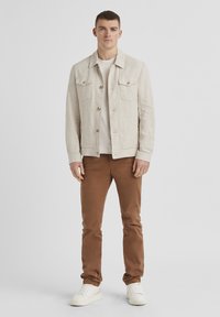 Beige Leinenjacke mit zwei Brusttaschen, kombiniert mit braunen Slim-Fit-Hosen und weißen Sneakers. Schlichtes Design ohne Muster oder Akzente.