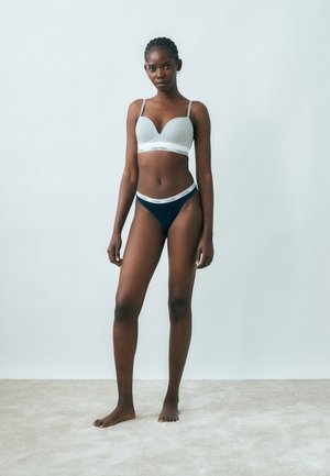 Donna con capelli intrecciati in piedi a piedi nudi, indossa un reggiseno grigio e mutande nere con fasce bianche di marca Calvin Klein, su uno sfondo chiaro semplice.