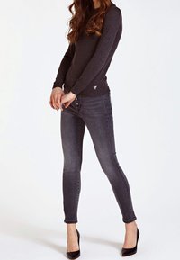 Donna che indossa un maglione grigio scuro aderente, jeans skinny neri scoloriti a vita alta e scarpe con tacco a punta nere, in piedi contro uno sfondo bianco.