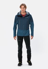 Heren blauwe waterdichte jas met capuchon, ritszakken, contrasterende donkere schouders, gecombineerd met zwarte technische broek en zwarte schoenen.