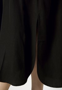 Jupe noire longueur genou avec une fente centrale au dos révélant une partie des jambes nues sur un fond uni.